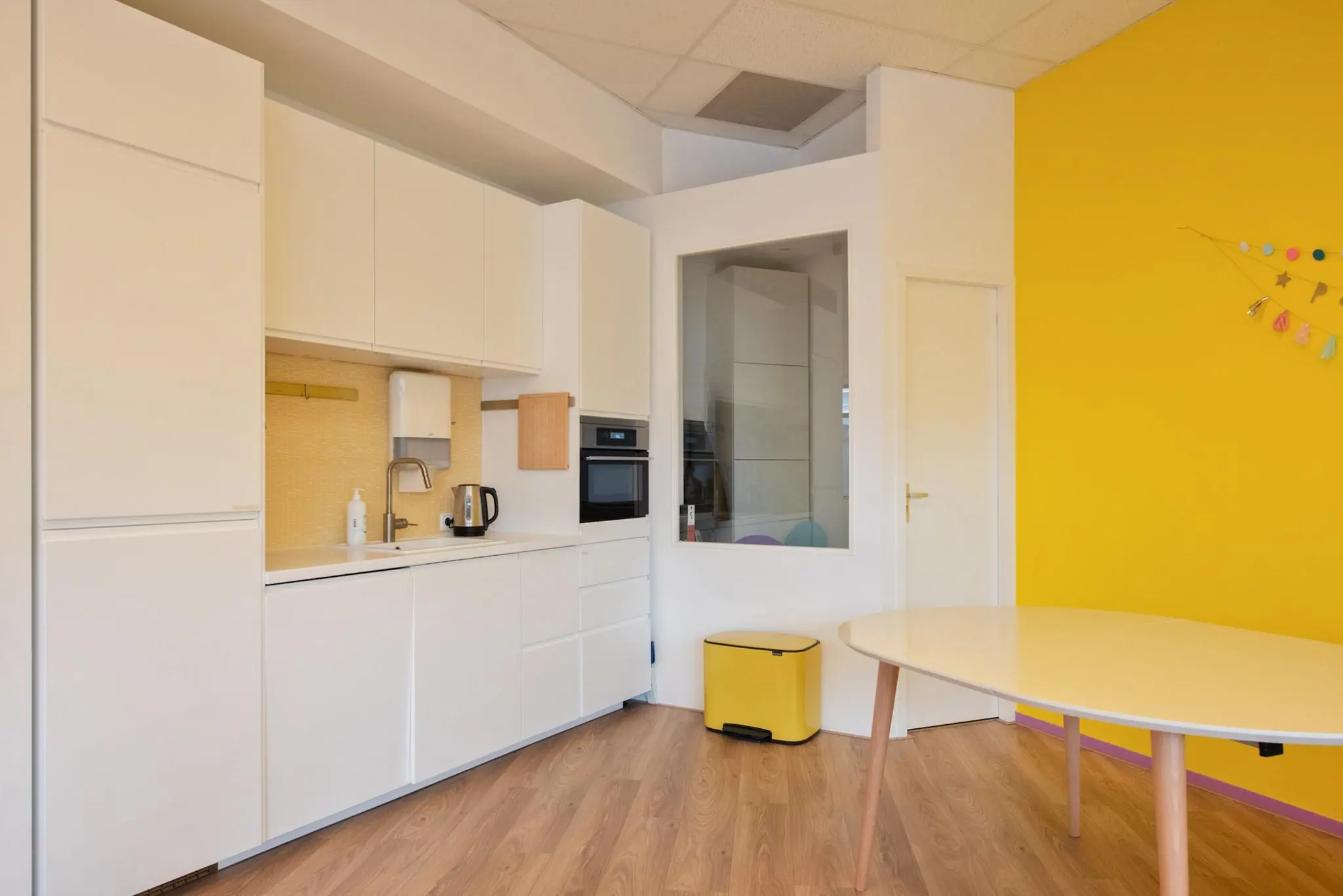 Moderne witte keuken met gele muur, houten vloer en een ovale eettafel in een ruimte aan de Koningin Julianalaan.